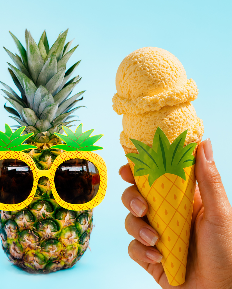 Hand holding a pineapple ice cream cone wrapped in a printable pineapple cone sleeve beside a fun pineapple wearing sunglasses on a bright tropical background.